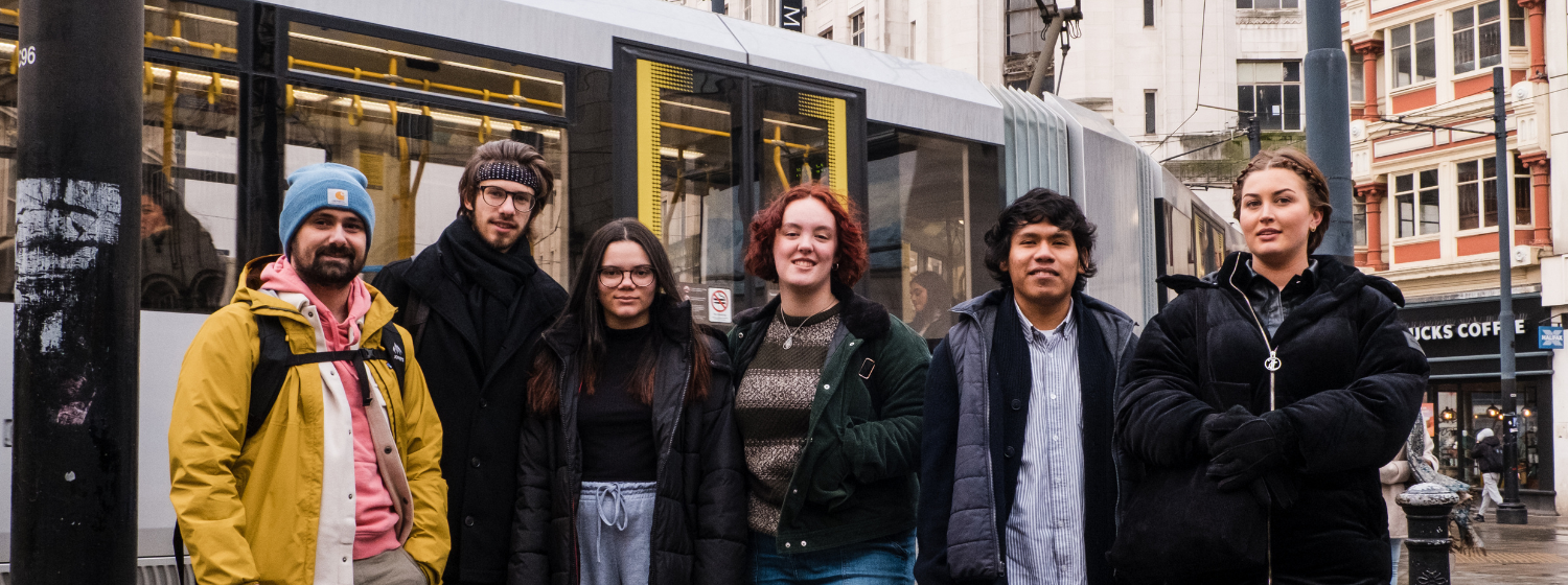 A group of students stands beside a passing tram in Manchester city centre.
