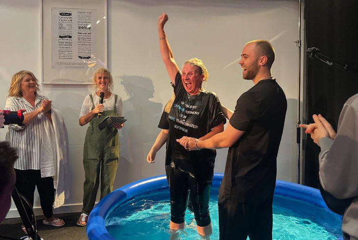 A woman punches the air with a look of joy on her face at her baptism. She stands in a baptism pool surrounded by friends.