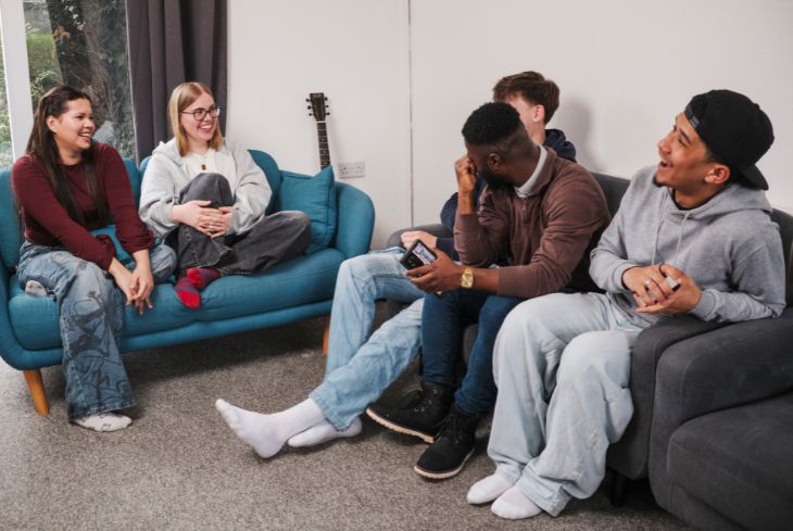 Five students sit around laughing in the sitting room of their accommodation.