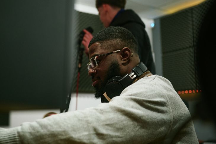 A student sits at a mixing desk in a recording studio with headphones around his neck.